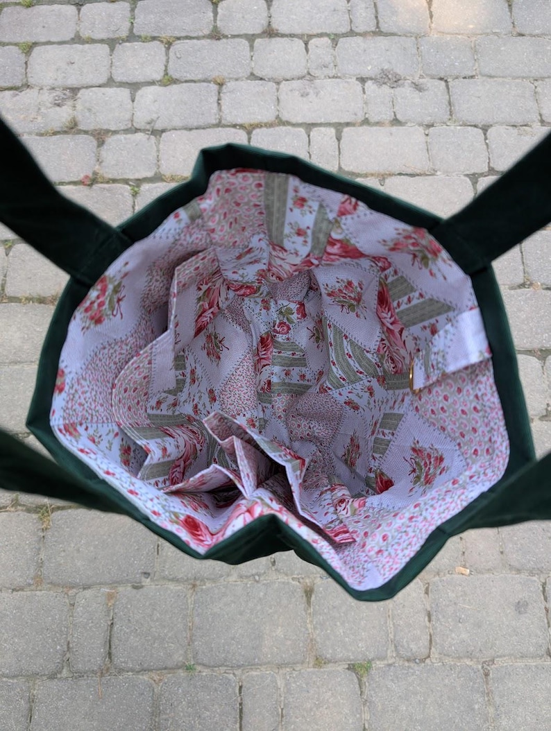 May include: A dark green tote bag with an open top, featuring a white interior lining decorated with a repeating pattern of pink roses, green leaves, and small floral sprigs. The bag includes multiple internal pockets along the sides.