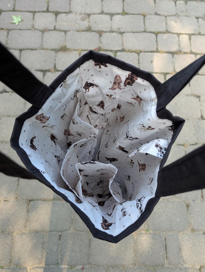 May include: A black tote bag with a white interior lining featuring a repeating pattern of brown and white spaniel dogs and small paw prints. The bag has two black fabric handles and internal pockets visible from an overhead perspective.