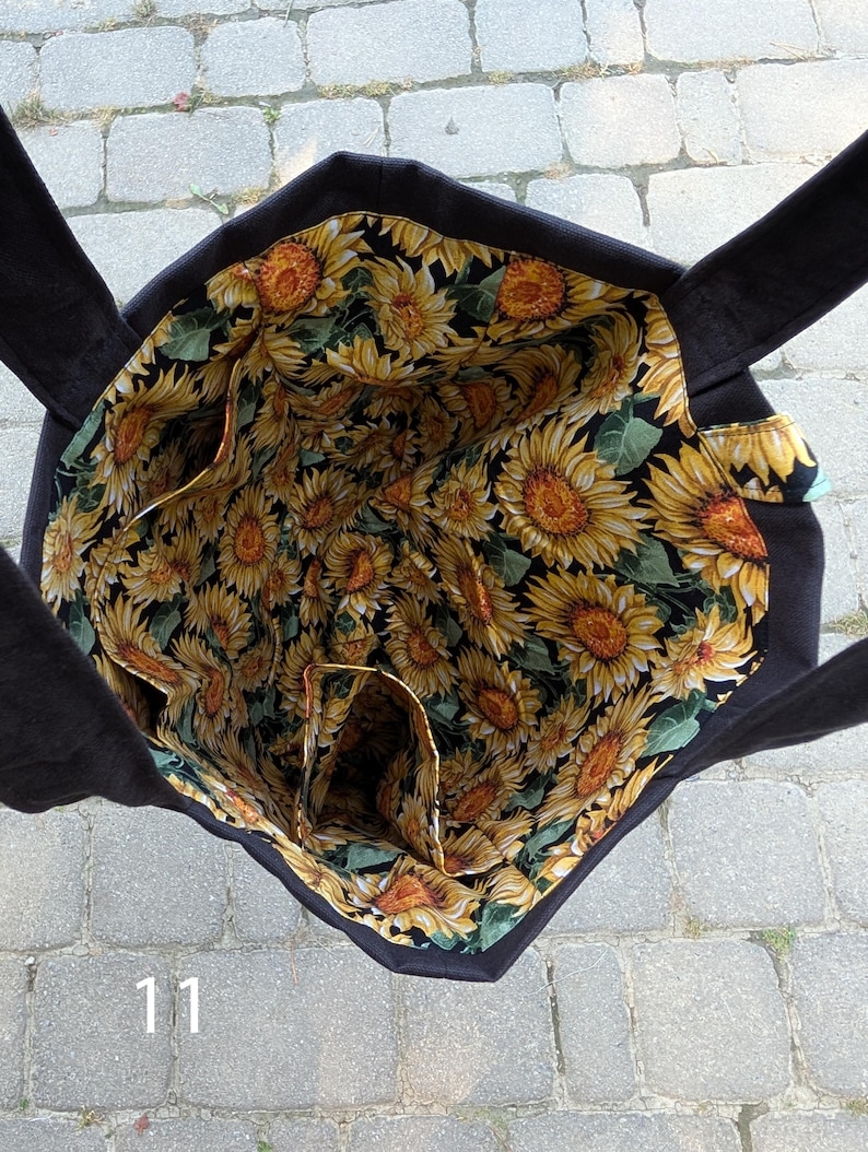 May include: A black tote bag with a sunflower print lining featuring yellow petals, brown centers, and green leaves on a black background. The interior includes multiple pockets and two black fabric handles extending from the top edges.