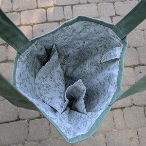 May include: A sage green tote bag with two shoulder straps, featuring an interior lining made of white fabric with a delicate grey floral line drawing pattern, including multiple internal pockets and a small metal D-ring attachment inside.