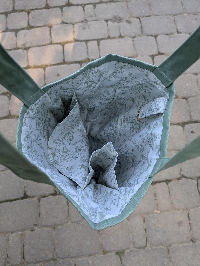 May include: A sage green tote bag with two shoulder straps, featuring an interior lining made of white fabric with a delicate grey floral line drawing pattern, including multiple internal pockets and a small metal D-ring attachment inside.