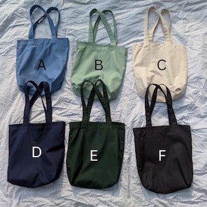 May include: Six solid-colored canvas tote bags arranged in two rows, labeled A through F. The top row features blue, sage green, and cream bags. The bottom row includes navy blue, dark forest green, and black bags, each with two shoulder straps.