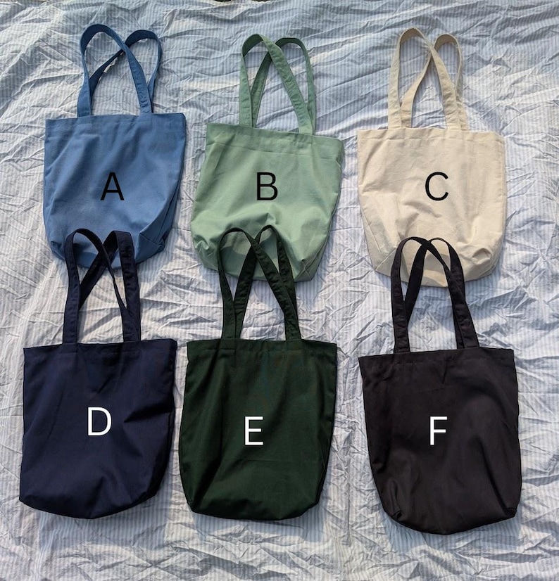 May include: Six solid-colored canvas tote bags arranged in two rows, labeled A through F. The top row features blue, sage green, and cream bags. The bottom row includes navy blue, dark forest green, and black bags, each with two shoulder straps.
