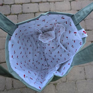 May include: A sage green tote bag with an open top and two shoulder straps. The interior is lined with white fabric featuring a repeating pattern of small red cherries and tiny black dots. The bag includes an internal pocket compartment.