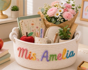 May include: A cream-coloured woven basket with the name "Miss Amelia" in rainbow yarn. Inside are flowers, a notebook, pens, a mug, and a small plant. A red apple and a pencil are also visible. A thoughtful gift.