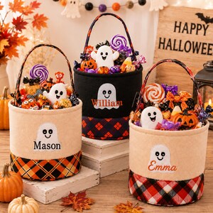 May include: Three cylindrical felt Halloween candy buckets with handles, each featuring a white ghost appliqué and a personalized name embroidered in orange or black. The buckets are filled with assorted candies and small pumpkin decorations.