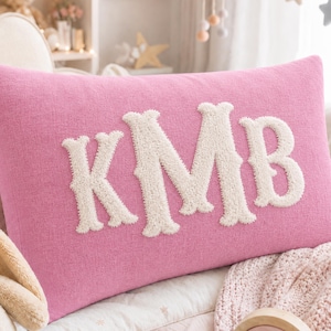Peut inclure: Un coussin rectangulaire rose orné des grandes lettres KMB brodées en relief de couleur crème au centre. Le coussin présente une texture en tissu tissé et est placé parmi d'autres objets doux dans un environnement de chambre d'enfant.