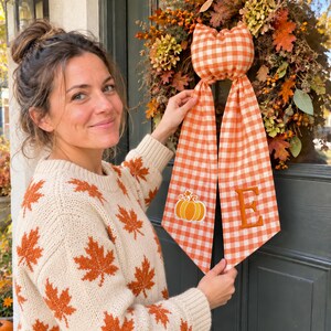May include: A fabric door hanger featuring an orange and white gingham pattern with a stuffed pumpkin-shaped top and two long tails. One tail displays an embroidered orange pumpkin, while the other features the embroidered capital letter E.