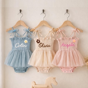 Könnte beinhalten: Drei Baby-Strampler in Blau, Beige und Rosa, jeweils mit Tüllrock und den Namen "Colin", "Olivia" und "Angela" auf dem Oberteil gestickt. Die Strampler hängen an Holzbügeln mit Schleifenriemen.