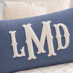 May include: A rectangular blue throw pillow featuring large, white, textured, embroidered letters spelling LMD in the center. The pillow has a soft, fabric-like surface and is positioned on a white quilted bedding surface alongside a teddy bear.