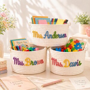May include: Three round white woven rope storage baskets with handles, each personalised with colourful embroidered names: Mrs. Anderson, Mrs. Brown, and Mrs. Davis. The baskets are filled with various school supplies, including colourful books, markers, and blocks.