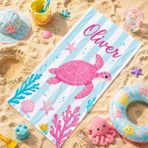 May include: A rectangular beach towel featuring light blue and white vertical stripes, a large pink sea turtle illustration, pink coral, and seashells. The name Oliver is printed in pink script text at the top of the towel, which is surrounded by beach toys.