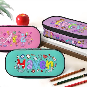 Puede incluir: Tres estuches personalizados con los nombres "Aria", "Lucy" y "Haven". El estuche rosa muestra "Aria", el morado "Lucy" y el turquesa "Haven". Cada uno tiene una cremallera negra e ilustraciones coloridas. Una manzana roja está al lado.