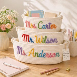 May include: A set of three stacked off-white woven rope baskets with handles. Each basket features colourful embroidered text: Mrs. Carter, Mr. Wilson, and Ms. Anderson. A small white tag attached to the bottom basket reads, Thank you for making a difference every day.