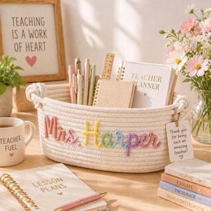 May include: An oval-shaped white cotton rope basket featuring the name Mrs. Harper embroidered in colourful yarn on the front. The basket contains various pens, a wooden ruler, and two notebooks. A white gift tag attached to the side reads Thank you for being such an amazing teacher!