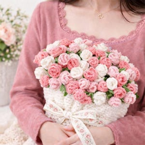 May include: A handcrafted bouquet of crochet roses in shades of pink and white, arranged in a white knitted basket. The basket is tied with a floral ribbon. The overall theme is a handmade floral arrangement.