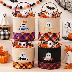 May include: Four cylindrical Halloween treat buckets with handles, featuring embroidered ghost designs and personalized names Lucas, Oliver, Henry, and William. The buckets have plaid fabric bases in purple, orange, and red, filled with assorted candies.
