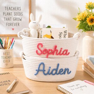 May include: Two stacked off-white woven rope baskets with integrated handles. The top basket features the name Sophia in pink cursive yarn lettering, and the bottom basket features the name Aiden in blue cursive yarn lettering.