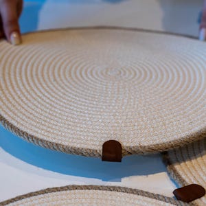 Puede incluir: Juegos de manteles individuales circulares tejidos, de color beige natural, con borde de cuerda marrón. Cada mantel individual tiene un pequeño detalle de cuero marrón. Dispuestos sobre una superficie blanca.
