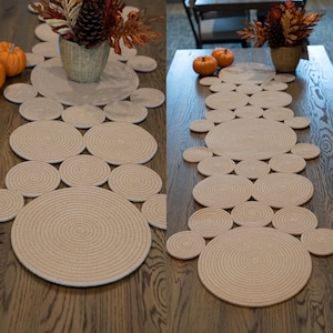 Può includere: Runner da tavolo in corda beige con un design circolare. Il runner è realizzato in corda avvolta strettamente, creando una superficie strutturata. Il design include cerchi grandi e piccoli, creando un motivo decorativo. Il runner è esposto su un tavolo di legno scuro.