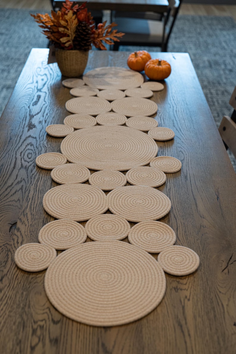 Pu&ograve; includere: Runner da tavolo beige in corda con un design circolare, posto su un tavolo in legno scuro. Il runner &egrave; circondato da sottobicchieri abbinati. Anche zucche decorative e una composizione floreale autunnale sono sul tavolo.