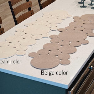 Könnte beinhalten: Zwei Tischläufer in Creme und Beige, aus gewebten, kreisförmigen Elementen. Der cremefarbene Läufer befindet sich links, der beige rechts. Die Texte "Cream color" und "Beige color" sind sichtbar.
