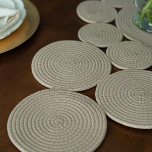 Pu&ograve; includere: Una collezione di sottobicchieri rotondi beige in corda, disposti su una superficie di legno marrone scuro. I sottobicchieri presentano un motivo a spirale. Un piatto dorato con piatti bianchi e un vaso di vetro sullo sfondo.