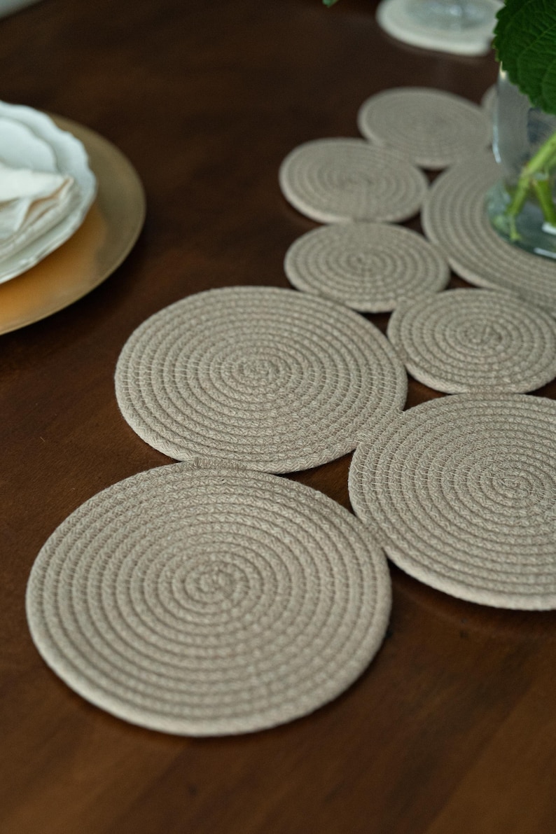 Pu&ograve; includere: Una collezione di sottobicchieri rotondi beige in corda, disposti su una superficie di legno marrone scuro. I sottobicchieri presentano un motivo a spirale. Un piatto dorato con piatti bianchi e un vaso di vetro sullo sfondo.