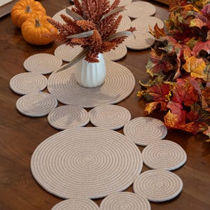 Pu&ograve; includere: Un'apparecchiatura da tavola decorativa con un runner e sottobicchieri in corda beige. Il tavolo &egrave; adornato con zucche arancioni, un vaso bianco con fogliame arancione e colorate foglie autunnali.