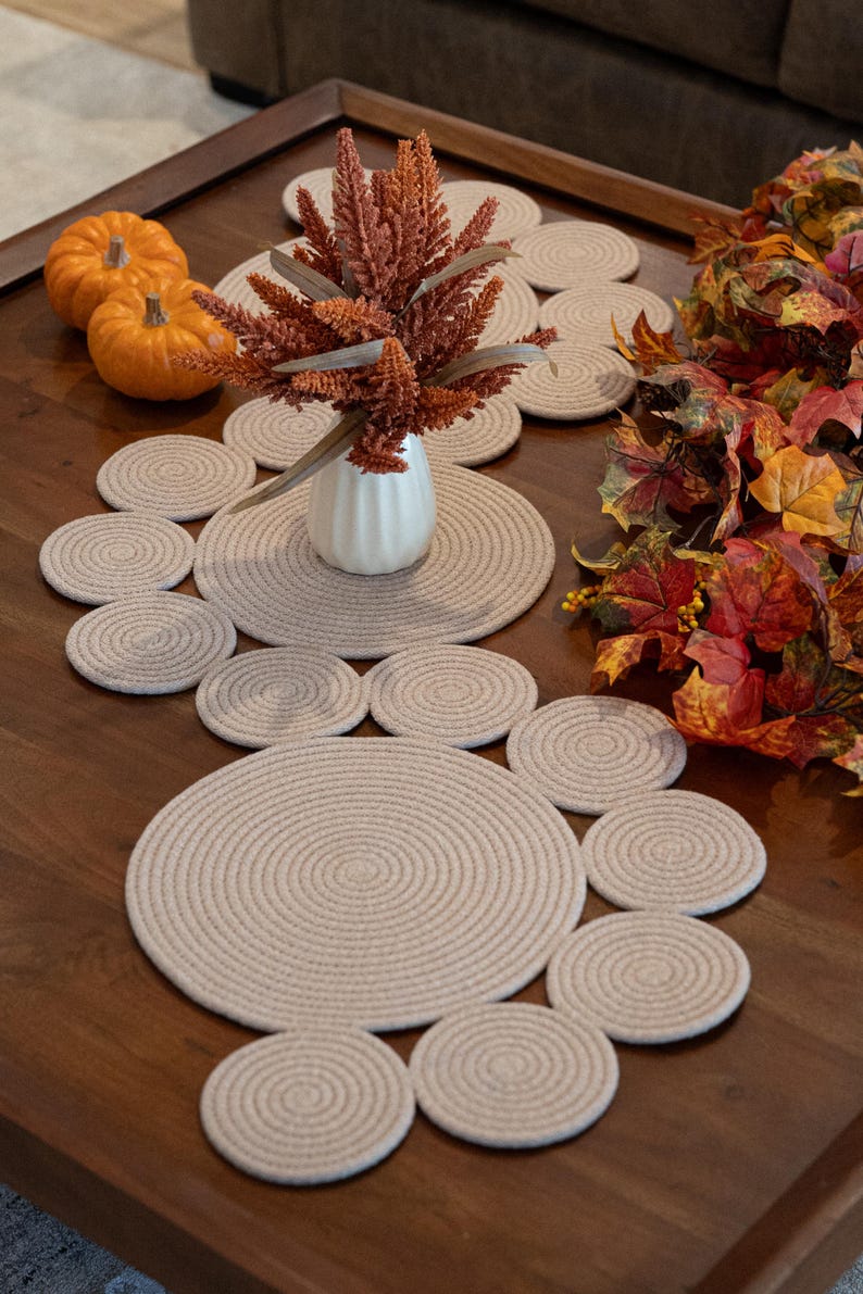 Pu&ograve; includere: Un'apparecchiatura da tavola decorativa con un runner e sottobicchieri in corda beige. Il tavolo &egrave; adornato con zucche arancioni, un vaso bianco con fogliame arancione e colorate foglie autunnali.