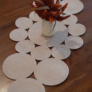 Pu&ograve; includere: Un'apparecchiatura da tavola decorativa con sottobicchieri in corda beige e un runner abbinato. Un piccolo vaso bianco contiene fiori arancioni secchi. I sottobicchieri e il runner sono disposti su un tavolo di legno marrone scuro.
