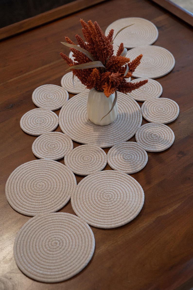 Pu&ograve; includere: Un'apparecchiatura da tavola decorativa con sottobicchieri in corda beige e un runner abbinato. Un piccolo vaso bianco contiene fiori arancioni secchi. I sottobicchieri e il runner sono disposti su un tavolo di legno marrone scuro.