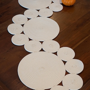 Puede incluir: Juegos de manteles individuales y posavasos de cuerda color crema dispuestos sobre una bandeja de madera oscura. Los manteles individuales son grandes, circulares y están rodeados de posavasos más pequeños. Dos pequeñas calabazas naranjas están en la bandeja.