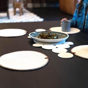 Pu&ograve; includere: Un tavolo da pranzo apparecchiato con tovagliette e sottobicchieri bianchi intrecciati. Una ciotola di cibo &egrave; al centro, circondata da tovagliette e sottobicchieri. Due tazze di ceramica grigie sono sul tavolo.