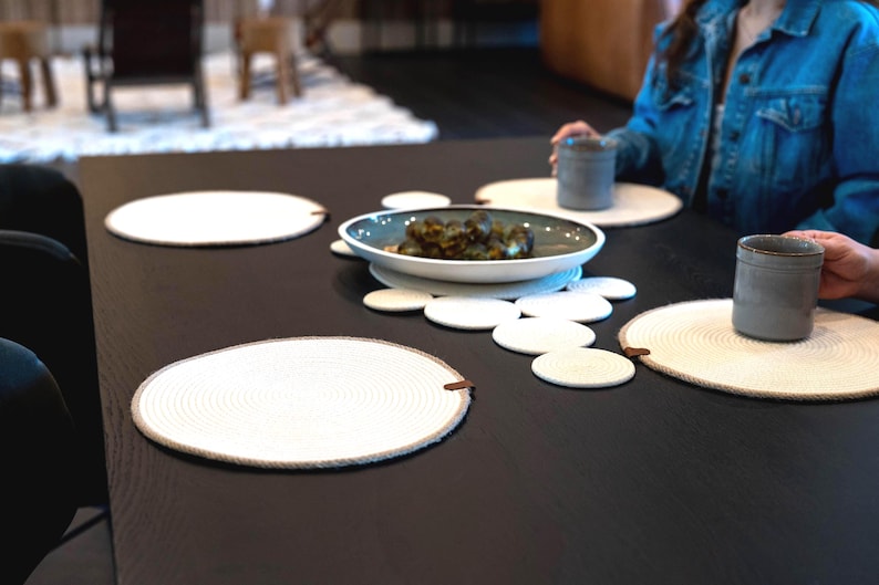 Pu&ograve; includere: Un tavolo da pranzo apparecchiato con tovagliette e sottobicchieri bianchi intrecciati. Una ciotola di cibo &egrave; al centro, circondata da tovagliette e sottobicchieri. Due tazze di ceramica grigie sono sul tavolo.