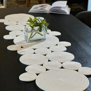 Pode incluir: Um caminho de mesa creme com um design circular, feito de corda enrolada. O caminho de mesa é colocado sobre uma mesa de madeira escura, com um pequeno vaso de vidro contendo uma planta verde. Um livro aberto é visível ao fundo.