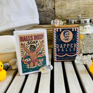 May include: Two boxes of novelty soap, one labeled "Balls Deep Soap" with a cartoon man in water, the other "Dapper Balls." Also, a yellow rubber duck, a small bathtub, and a glass jar with a silver lid are visible.