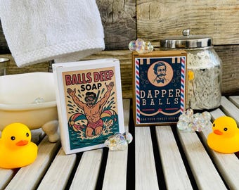 May include: Two novelty soap boxes, one reading "Balls Deep Soap" with a cartoon man in water, the other "Dapper Balls." A yellow rubber duck, a small bathtub, and a glass jar with a silver lid are also present.