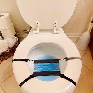 May include: White toilet with the lid open, featuring a black and blue mesh guard suspended within the bowl. The guard is secured by black straps attached to the toilet seat. A roll of toilet paper and a toilet brush are visible in the background.
