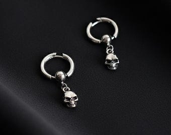 May include: A pair of silver-colored hoop earrings, each with a small skull charm dangling from the bottom. The earrings are set against a dark, textured background, creating a striking contrast.