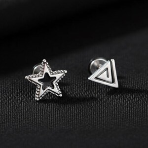 May include: A pair of silver stud earrings. One earring is a star shape with a black center and a dotted border. The other earring is a triangle shape with a spiral design. The earrings are displayed on a black surface.