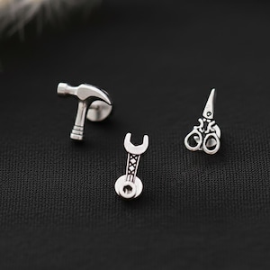 Könnte beinhalten: Silberne Ohrstecker mit den Motiven Hammer, Schraubenschlüssel und Schere. Die Ohrringe sind klein und detailliert, mit poliertem Finish. Der Hammer hat einen strukturierten Kopf, der Schraubenschlüssel ein detailliertes Design und die Schere offene Klingen. Auf schwarzem Hintergrund.