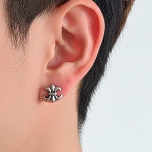 May include: A silver-colored stud earring in the shape of a fleur-de-lis is worn on the ear. The earring is a small, detailed design with three petals and a central element. The earring is set against the backdrop of a person's ear and neck.