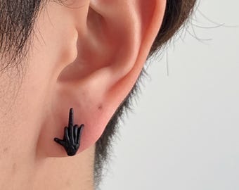 Puede incluir: Pendiente de botón negro con forma de mano con el dedo corazón levantado. El pendiente se lleva en la oreja de una persona con cabello oscuro. El fondo es blanco liso.