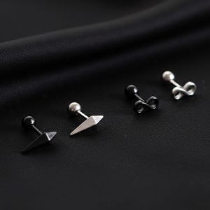 Op de afbeelding: Vier stud oorbellen in zilver en zwart, gepresenteerd op zwarte stof. De oorbellen hebben geometrische vormen, waaronder een driehoek, een oneindigheidssymbool en een kogelstud. De kleine, delicate oorbellen zijn geschikt voor dagelijks gebruik.