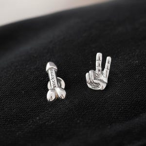 Puede incluir: Un par de pendientes plateados. Uno tiene forma de pene con la palabra "suckme" grabada. El otro es una mano haciendo el signo de la paz. Los pendientes se muestran sobre un fondo negro.