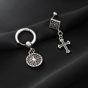 Puede incluir: Un par de pendientes plateados. Uno es un aro con un dije circular. El otro es un perno con un dije de cruz. Los pendientes se muestran sobre una superficie negra.