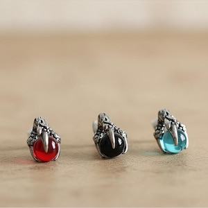 May include: Three silver claw earrings, each holding a coloured orb: red, black, and blue. The earrings have a detailed, textured design, and the orbs are smooth and spherical. The background is a blurred, neutral tone.