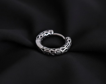 May include: A silver hoop earring with a detailed, repeating pattern. The earring has a hinged clasp. The design features a series of stylized shapes and lines, creating a textured appearance. The earring is set against a dark background.
