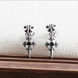 Peut inclure: Une paire de boucles d'oreilles argentées avec un motif de croix gothique. Chaque boucle d'oreille présente un dessus en fleur de lys, une pierre noire centrale et un bas pointu. Les boucles d'oreilles sont présentées sur un fond clair.